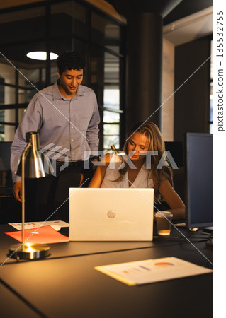 Colleagues collaborating on laptop in modern office during late-night work session Colleagues collaborating on laptop in modern office during late-night work session 135532755