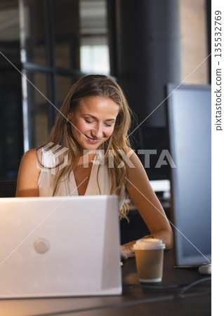 Businesswoman working late at office, smiling while using laptop with coffee nearby Businesswoman working late at office, smiling while using laptop with coffee nearby 135532769