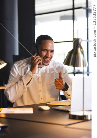 Smiling man giving thumbs up while talking on phone at office desk Smiling man giving thumbs up while talking on phone at office desk 135532777