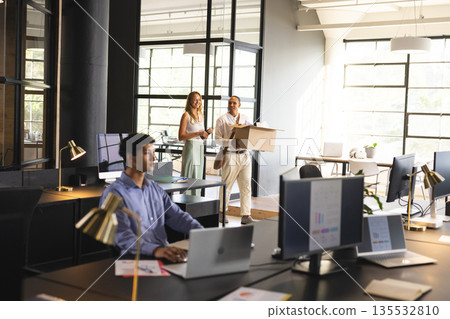 Colleagues carrying box in modern office while coworker working on laptop Colleagues carrying box in modern office while coworker working on laptop 135532810