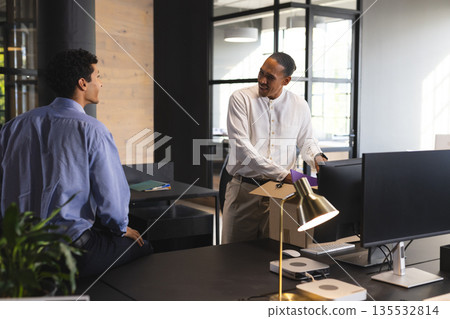 African American businessman unpacking box at office desk, talking with colleague African American businessman unpacking box at office desk, talking with colleague 135532814
