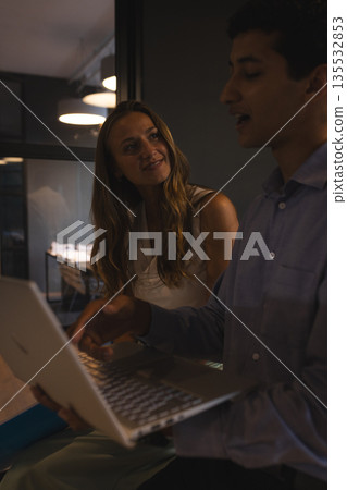 Colleagues discussing project while using laptop in modern office at night 135532853