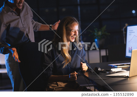 Smiling woman celebrating success at office desk with colleague at night Smiling woman celebrating success at office desk with colleague at night 135532890