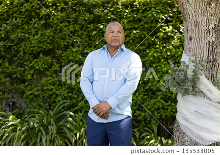 Senior man standing outdoors by tree, looking thoughtful in garden setting Senior man standing outdoors by tree, looking thoughtful in garden setting 135533005