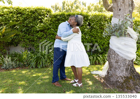 Senior couple embracing lovingly in sunny garden near tree with white ribbon Senior couple embracing lovingly in sunny garden near tree with white ribbon 135533040
