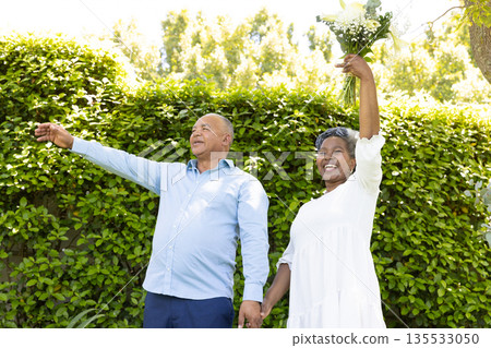 Senior couple celebrating outdoors, holding hands and raising bouquet joyfully 135533050