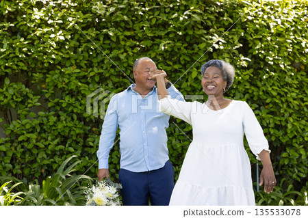 Senior couple dancing joyfully in garden, holding hands and smiling brightly 135533078