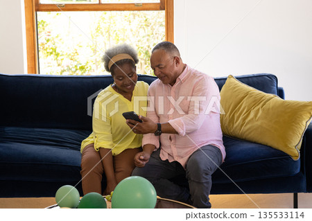 Senior couple sitting on sofa, smiling while looking at smartphone together Senior couple sitting on sofa, smiling while looking at smartphone together 135533114