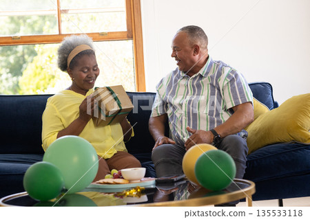 Senior couple celebrating at home, woman opening gift with joyful expression Senior couple celebrating at home, woman opening gift with joyful expression 135533118