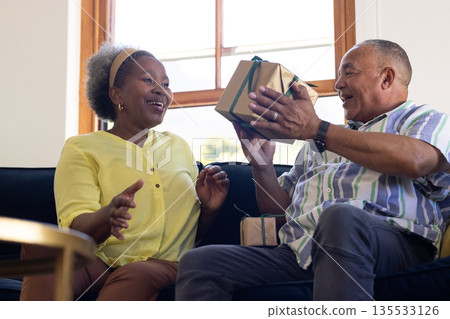 African American senior couple exchanging gift on couch, both looking joyful 135533126