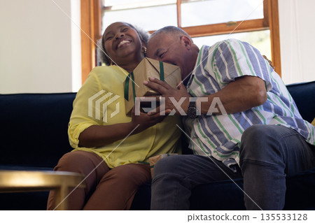 Senior couple joyfully embracing while holding gift on living room sofa Senior couple joyfully embracing while holding gift on living room sofa 135533128