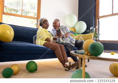 African American senior couple sitting on sofa, smiling and looking at smartphone African American senior couple sitting on sofa, smiling and looking at smartphone 135533129