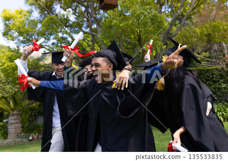 Graduating teens celebrating outdoors, wearing caps and gowns, holding diplomas 135533835