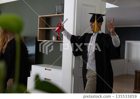 Teen celebrating graduation at home, wearing cap and gown, holding diploma Teen celebrating graduation at home, wearing cap and gown, holding diploma 135533859