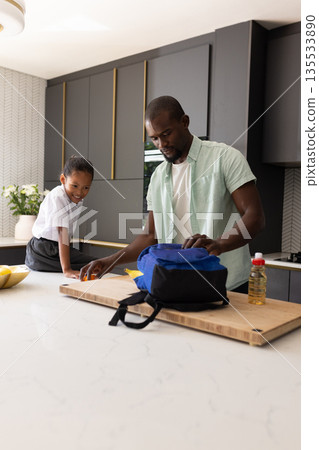 African American father packing lunch while child watches in modern kitchen 135533890