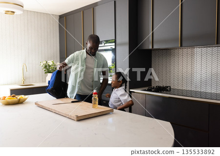 Father helping daughter pack backpack in modern kitchen before school Father helping daughter pack backpack in modern kitchen before school 135533892