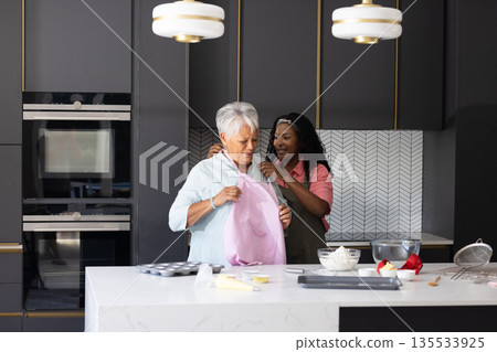 Granddaughter helping grandmother put on apron in modern kitchen, preparing to bake Granddaughter helping grandmother put on apron in modern kitchen, preparing to bake 135533925
