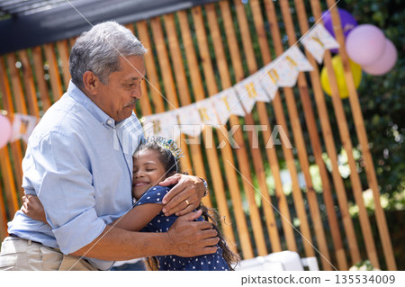 Grandfather hugging granddaughter at outdoor birthday party, both smiling warmly 135534009