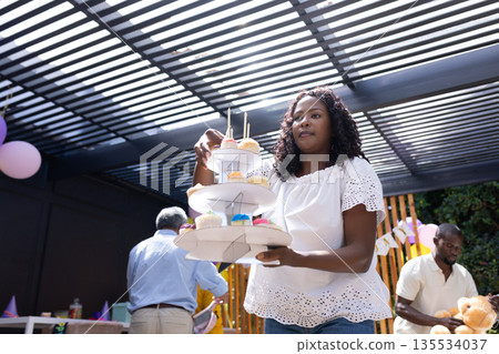 African American woman arranging colorful cupcakes on tiered stand at family party African American woman arranging colorful cupcakes on tiered stand at family party 135534037