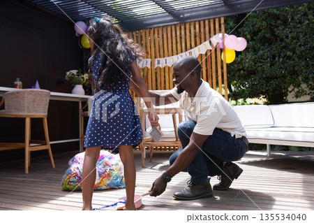 Helping daughter open birthday gift, African American father smiling outdoors together 135534040