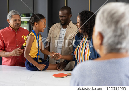 Proud family celebrating young girl with trophy and medal in kitchen 135534063