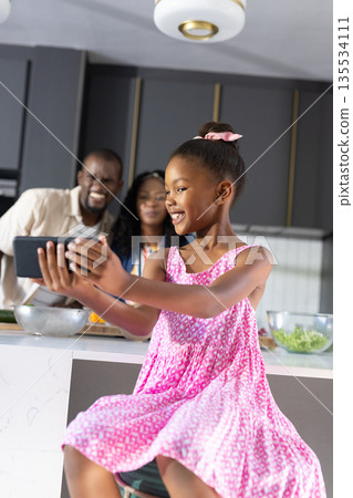 Young girl in pink dress taking selfie with family in modern kitchen 135534111