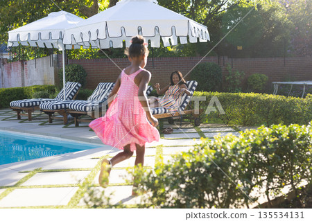 Young girl in pink dress running by pool while woman relaxes on lounge chair Young girl in pink dress running by pool while woman relaxes on lounge chair 135534131