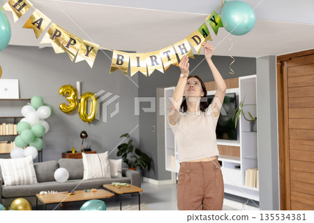 Woman hanging HAPPY BIRTHDAY banner in living room with gold 30 balloon and mint-green balloons 135534381