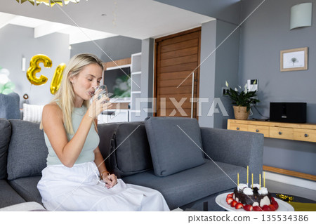 Woman in twenties drinking water on sofa in living room, with chocolate cake, gold number balloons 135534386