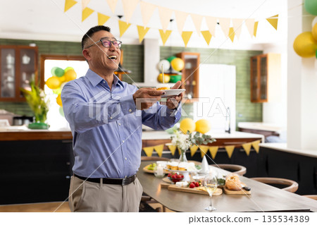 Senior Chinese Asian man standing and holding plate with lit cupcake in festive kitchen, copy space Senior Chinese Asian man standing and holding plate with lit cupcake in festive kitchen, copy space 135534389