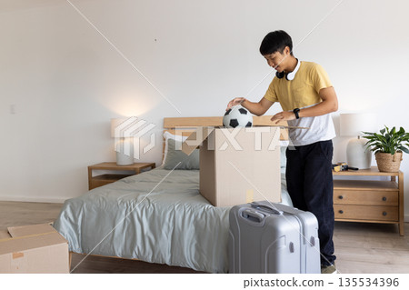 Asian man unpacking soccer ball from cardboard box in bedroom with rolling suitcase, copy space Asian man unpacking soccer ball from cardboard box in bedroom with rolling suitcase, copy space 135534396