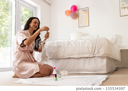 Woman painting nails on towel next to bed in bedroom, with nail polish bottles, nail brush 135534397
