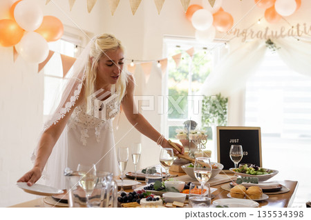 Bride wearing lace gown and veil arranging dishes on table in indoor reception with peach balloons 135534398