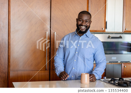 African American man leaning on white kitchen island in modern kitchen, holding tan ceramic mug African American man leaning on white kitchen island in modern kitchen, holding tan ceramic mug 135534400