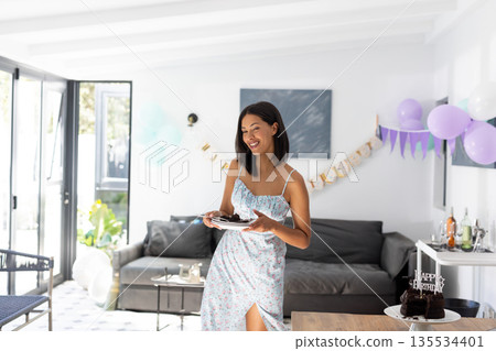 Woman holding cake slice plate and celebrating birthday in modern living room, with pastel balloons 135534401