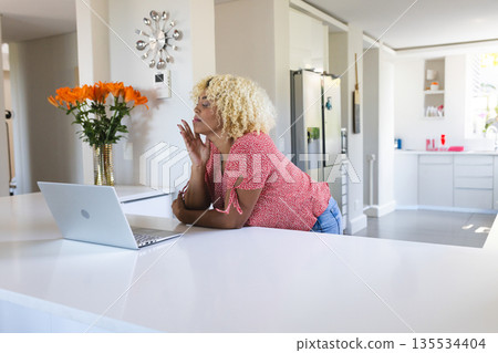 Leaning on kitchen island wearing red blouse and jeans, gazing at silver laptop and orange lilies Leaning on kitchen island wearing red blouse and jeans, gazing at silver laptop and orange lilies 135534404