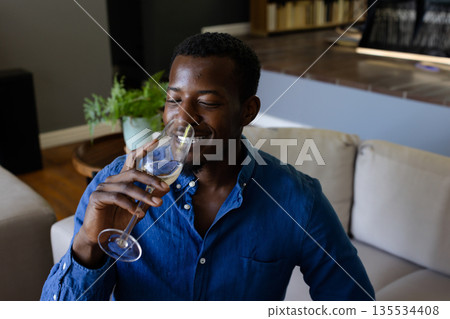 African American man drinking white wine from glass on beige couch in modern living room African American man drinking white wine from glass on beige couch in modern living room 135534408