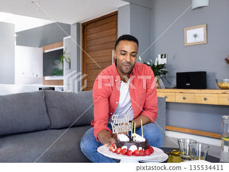 African American man holding birthday cake with lit candles on couch in living room, smiling 135534411