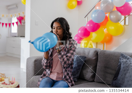 Woman inflating blue balloon on gray couch in decorated living room, with birthday cake and bunting Woman inflating blue balloon on gray couch in decorated living room, with birthday cake and bunting 135534414