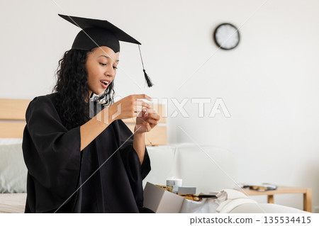 Teenage girl opening gift box wearing graduation gown with mortarboard on bed, examining necklace Teenage girl opening gift box wearing graduation gown with mortarboard on bed, examining necklace 135534415