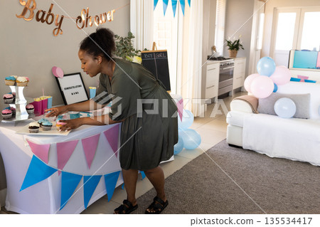 African American pregnant woman arranging cupcakes in living room, with baby shower banner 135534417