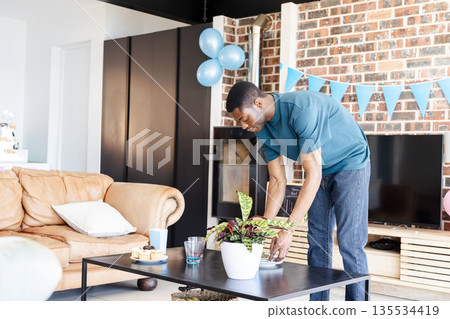 Mid adult African American man arranging potted plant on coffee table in living room, copy space 135534419