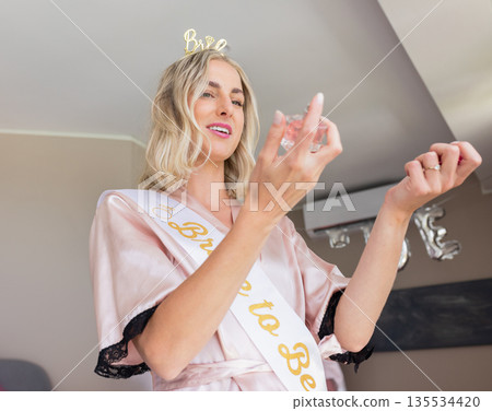 Woman standing in party room, wearing pink satin robe and sash, holding clear glass and smiling Woman standing in party room, wearing pink satin robe and sash, holding clear glass and smiling 135534420