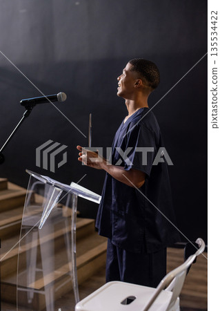 Man standing behind podium with microphone on stage holding trophy smiling upward, copy space Man standing behind podium with microphone on stage holding trophy smiling upward, copy space 135534422