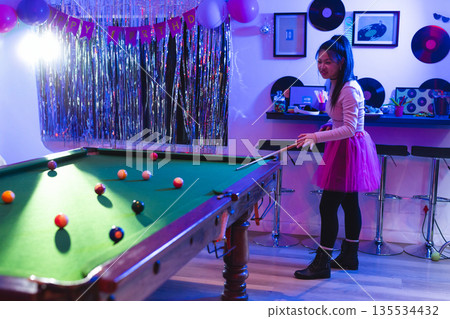 Young woman playing on pool table in home party room, with birthday decorations, copy space 135534432
