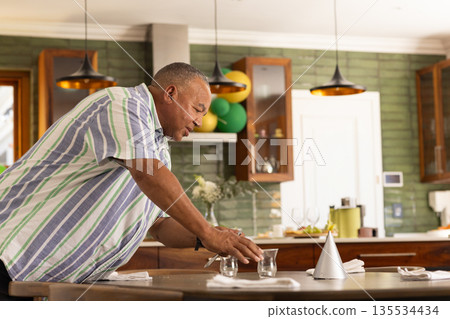 Senior man placing party hat and glasses on dining table in kitchen with balloons, copy space 135534434