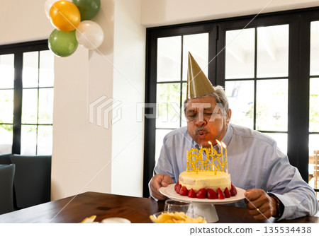 Mid adult man celebrating 60th birthday at home dining table with cake, party hat, copy space 135534438