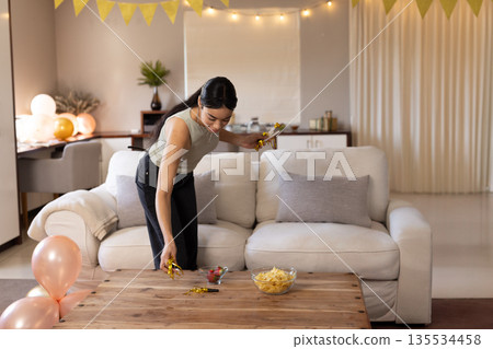 young woman placing golden party poppers on coffee table in living room with balloons, copy space young woman placing golden party poppers on coffee table in living room with balloons, copy space 135534458