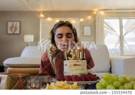 Female leaning forward and blowing out cake candles in living room, with gift, chips and grapes Female leaning forward and blowing out cake candles in living room, with gift, chips and grapes 135534470
