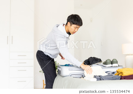 Asian man packing silver suitcase on bed in minimalist bedroom at home, with clothes and sneakers 135534474